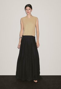 Beige ärmelloses Top mit strukturierter Form und subtiler Textur, kombiniert mit einem fließenden schwarzen Maxikleid. Model trägt ein silbernes Armreif.