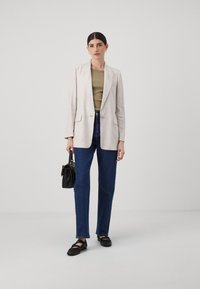 Blazer beige chiaro, top verde oliva, jeans blu scuro, mocassini neri, in mano una piccola borsa nera, in posizione su uno sfondo neutro.