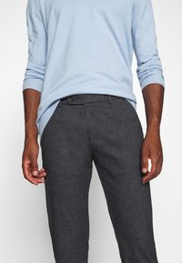 Mężczyzna w jasnoniebieskim swetrze z długim rękawem wsuniętym w ciemnoszare dopasowane spodnie z guzikami i szlufkami na pasek. Ręce swobodnie opadają wzdłuż ciała.