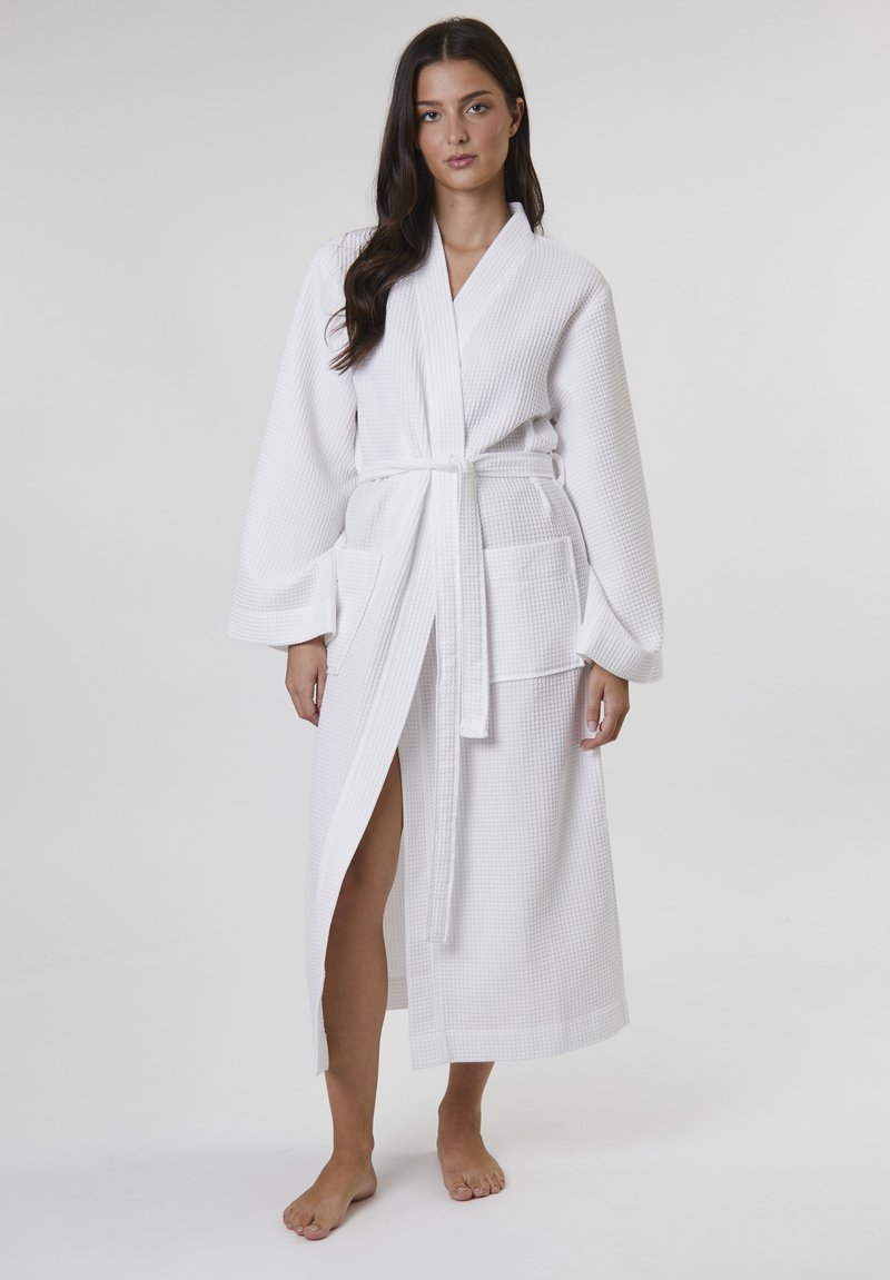 Accappatoio bianco in maglia waffle con maniche lunghe, cintura da annodare e due tasche frontali. Presenta un'apertura laterale per facilitare i movimenti.