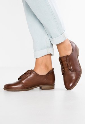 Brązowe skórzane buty oxfordy charakteryzują się niskim obcasem, detalami sznurowania i teksturowanym wzorem. Noszone z podwiniętymi jasnoniebieskimi dżinsami.