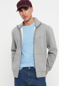 Sudadera gris con cremallera y cuello alto, que presenta acentos en rojo y azul en contraste. Usada sobre una camiseta de color azul claro. Diseño sencillo.