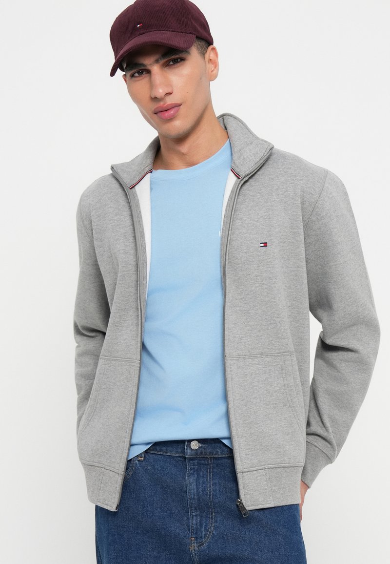 Sudadera gris con cremallera y cuello alto, que presenta acentos en rojo y azul en contraste. Usada sobre una camiseta de color azul claro. Diseño sencillo.