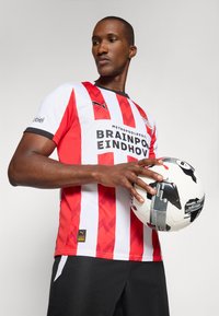 Rød og hvid stribet fodboldtrøje med sort krave og ærmer, prydet med et logo og tekst. Spilleren holder en sort og hvid mønstret fodbold.