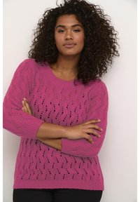 Maglione rosa lavorato a maglia con una trama allentata, caratterizzato da un motivo a onda e colletto e polsini a coste. Il modello posa con le braccia incrociate.