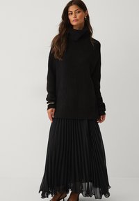 Sort strikket sweater med høj krave, parret med en lang, plisseret sort nederdel lavet af letvægtsmateriale. Elegant og flydende design.