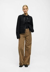 Černý pletený kardigan s mašlí, kombinovaný s hnědými kordovými kalhotami s širokými nohavicemi a černými lesklými botami. Hladká textura a uvolněný střih.