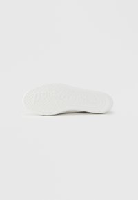 Semelle de sneaker blanche avec motif texturé, présentant des indentations circulaires et un logo embossé. Les côtés lisses s'effilent vers la pointe.