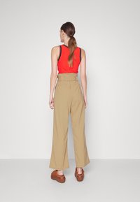 Rotes, geripptes, ärmelloses Oberteil mit blauen Akzenten, kombiniert mit hochtaillierten beigen, weiten Hosen. Braune Plateau-Schuhe runden das Outfit ab.