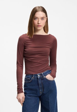 Frau mit glatten hellbraunen Haaren, die ein braunes, eng anliegendes Langarmshirt und hoch taillierte blaue Jeans trägt, mit einer Hand in der Tasche.
