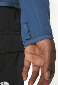 Niebieski rękaw z polaru z zapięciem na guziki, pokazujący teksturowaną tkaninę. Czarne ubranie widoczne poniżej, kontrastujące kolorem i materiałem.