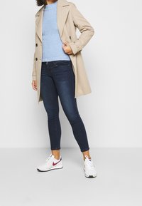 Beige trenchcoat, gestructureerde lichtblauwe trui, donkerblauwe skinny jeans en witte sneakers met rode accenten. Schoon ontwerp, nauwsluitende pasvorm.