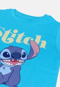 Ljusblå skjorta med en leende lila tecknad figur, Stitch, och stor beige text som säger "Stitch" på framsidan.