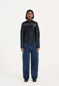 Svart skinnjacka med hög krage, vita ränder längs axlarna och broderade detaljer på bröstet, kombinerad med oversized blå jeans.