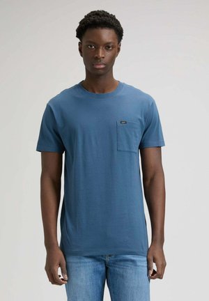 Giovane uomo che indossa una t-shirt blu a maniche corte con una tasca sul petto e jeans azzurri, in piedi davanti a uno sfondo semplice.