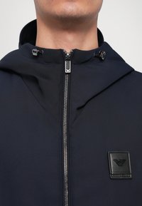 Tamnoplava jakna na patentni zatvarač s kapuljačom, izrađena od teksturiranog materijala s istaknutom crnom logotip oznakom na prsima. Srebrni patentni zatvarač.