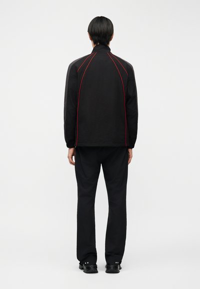 Άτομο που στέκεται με την πλάτη γυρισμένη, φορώντας μαύρο μπουφάν με κόκκινες ραφές, μαύρο παντελόνι και μαύρα παπούτσια σε απλό ανοιχτό φόντο.