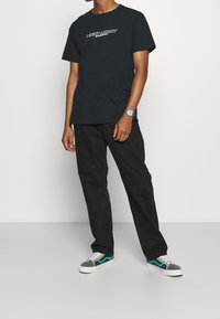 Černé bavlněné tričko s grafikou "NIGHT ADDICT", spárované s tmavými kalhotami a zelenošedými botami s bílými akcenty a tkaničkami.