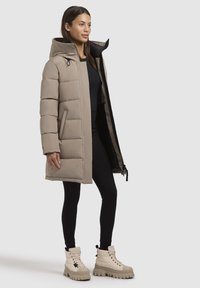 Parka bej căptușit cu glugă, fermoar frontal și buzunare laterale. Purtați peste un top negru și asortat cu cizme albe masive și colanți negri.