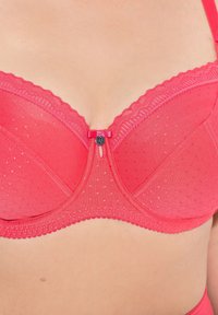 Röd bh med genomskinlig prickig design, underwire stöd, spetskant och en central rosett med metallisk detalj vid stängningen.