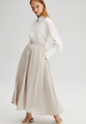 Camicia bianca a maniche lunghe abbinata a una lunga gonna beige con vita alta e bottoni frontali. Tessuto morbido con una silhouette fluida.