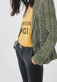 Grönmönstrad kavaj med vita mönster, gul v-ringad t-shirt med svart text och svarta byxor. Outfitten visar upp lager av texturer.