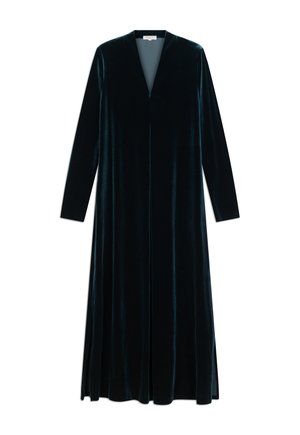 Personne aux cheveux courts portant une longue robe en velours bleu foncé, tenant un sac à main sombre et portant des bottes marron à talons hauts, vue de dos.