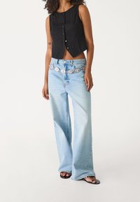 Czarny, bezrękawnikowy top w kroju cropped z przednimi guzikami, połączony z jasnoniebieskimi spodniami o szerokich nogawkach, z dekoracyjnym, wielokolorowym paskiem.