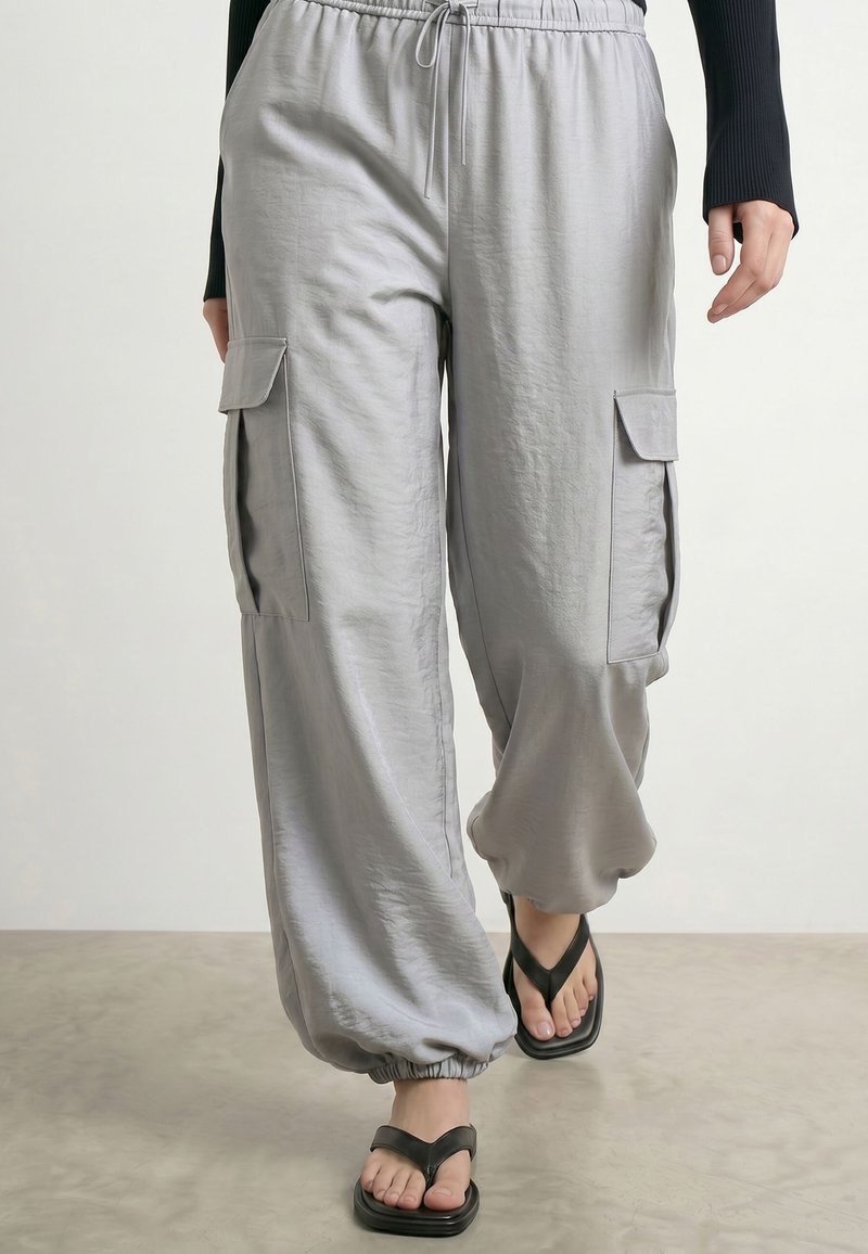 Personne portant un pantalon cargo gris clair ample avec des poignets élastiques et des tongs noires, marchant sur une surface plane avec un fond blanc.