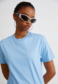 Maglietta di cotone azzurro chiaro con un discreto logo bianco sul lato sinistro del petto, abbinata a occhiali da sole con accenti argentati unici e orecchini a cerchio.