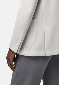 Blazer grigio chiaro con un tessuto textured, dotato di una tasca singola e tre bottoni decorativi sul polsino. Abbinato a un pantalone grigio scuro.