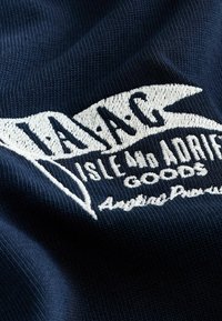 Tmavomodrá pletená tkanina s bielym vyšívaným textom „TAJAG ISLE AND ADRIATIC GOODS“ a menším kurzívnym písmom pod ním.