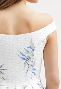 Robe blanche à épaules dénudées en tissu texturé, présentant un motif floral bleu et vert dans le dos, mettant en valeur des détails complexes.