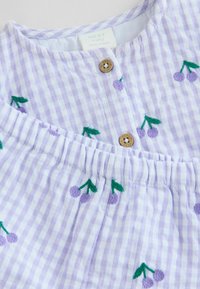 Tessuto a righe lilla e bianche con ciliegie viola ricamate e foglie verdi. Presenta una pattina con bottoni e un elastico in vita.