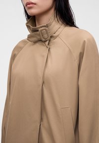 Femme portant un manteau beige à col montant avec une fermeture à boucle et sangle autour du cou, vue des épaules au menton.