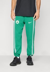 Green athletic pants with white stripes and Celtics logo. Made of smooth fabric, with elastic cuffs and pockets. Worn with black sneakers.