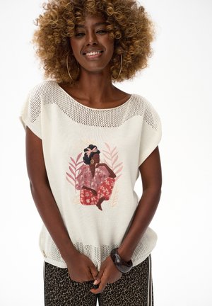 Vrouw met krullend haar, draagend een grote oorbel, een wit gebreid topje met een illustratie van een zittende vrouw, en een broek met patroon, glimlachend zachtjes.