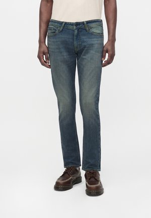 Man die vervaagde blauwe denim jeans en donkerbruine leren veterschoenen draagt, staand tegen een witte achtergrond.