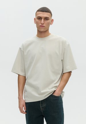 Jeune homme aux cheveux courts portant un T-shirt beige clair surdimensionné et un jean foncé, debout avec une main dans la poche sur un fond uni.