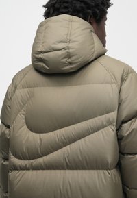 Prešita bunda v olivno zeleni barvi s kapuco. Ima prešito zasnovo, mehko teksturo in izrazit diagonalni vzorec šivov.