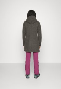 Cappotto lungo grigio scuro con cappuccio, caratterizzato da una silhouette aderente. Abbinato a pantaloni rosa brillante e scarpe nere con dettagli color turchese.