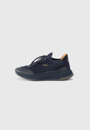 Navy blå BOSS sneaker med tekstureret strikket overdel, snørelukning, tyk rillede sål og tan trækflig på hælen, vist fra siden på en ensfarvet baggrund.