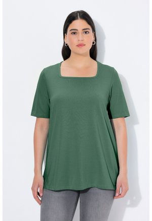 Vrouw met donker haar die een groene top met korte mouwen en een vierkante halslijn draagt en lichtgrijze spijkerbroek, staand tegen een effen witte achtergrond.