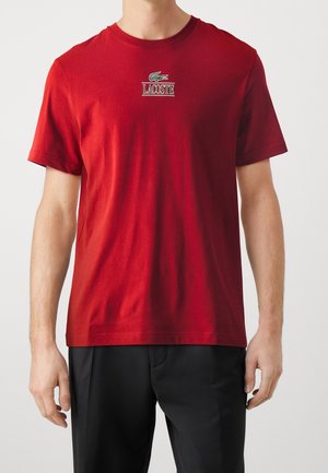 Homme portant un t-shirt rouge à manches courtes avec un petit logo crocodile vert et le texte "LACOSTE" sur la poitrine, associé à un pantalon noir.
