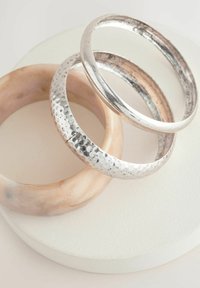 Trois bracelets sont exposés : un lisse et rose pâle, et deux en argent, l'un avec une texture martelée et l'autre poli, reposant sur un piédestal blanc.