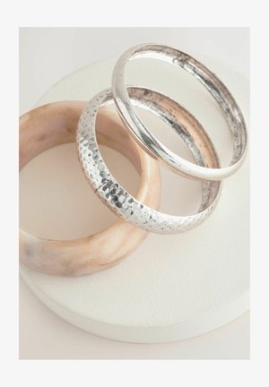 Trei brățări sunt expuse: una netedă, roz deschis, și două din argint, una cu o textură băltită și cealaltă lustruită, așezate pe un piedestal alb.