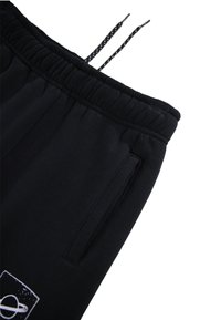 Pantalon de survêtement noir avec une taille élastique et des cordons de serrage, poches latérales et une patch logo sur la jambe gauche. Texture de tissu douce.
