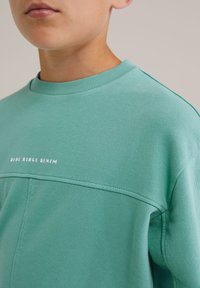 Artimas planas asmens, vilkinčio melsvai žalią džemperį su užrašu "BLUE RIDGE DENIM" baltąja spalva krūtinėje.