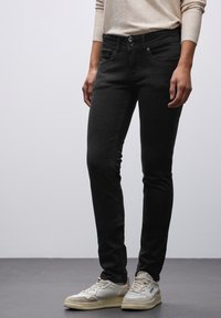 Jeans skinny neri con una texture liscia, cinque tasche e chiusura a bottone, abbinati a sneaker bianche e beige.