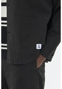 Black lightweight jacket with a textured finish, featuring a large patch pocket and a label with "M 73." Accents include visible stitching.